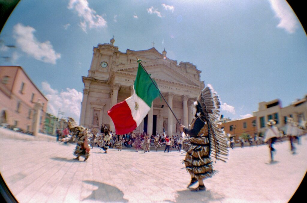 A colorful parade in Mexico with traditional costumes and a Mexican flag under a sunny sky.